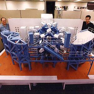 Laser fusion reactor scale model with blue steel framework, stairs and white beamline units, two men standing beside it