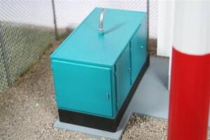 Teal standby generator on the cell tower model, sitting on its concrete pad beside the red and white monopole base