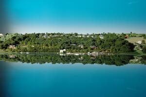 Lakefront site model seen at water level, wooded shoreline with homes and docked boats mirrored in the glossy lake