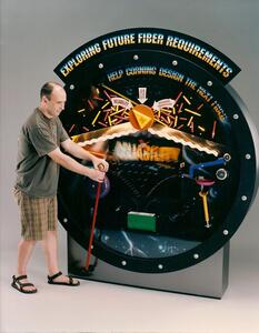 Giant pachinko machine model with working parts, a man pulling its red plunger lever under a fiber-themed header sign