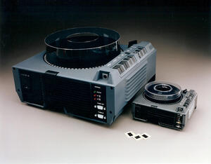 Oversized Kodak slide projector display model with clear carousel top beside a smaller projector and three 35mm slides