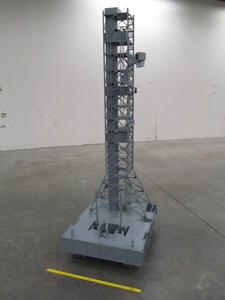Rocket launch pad model with tall gray lattice tower on a tracked base, shown on a warehouse floor beside a yardstick