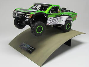Trophy truck model mounted mid-jump on a curved dirt-painted steel base with a nameplate, front three-quarter view