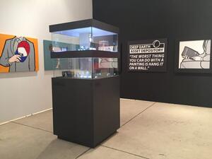 Art tower model in a two-level glass display cabinet on a dark pedestal, shown in a gallery beside pop art canvases