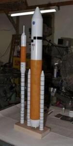 Ares I and Ares V rocket scale models side by side on a shared wooden base, showing orange core stage and twin boosters