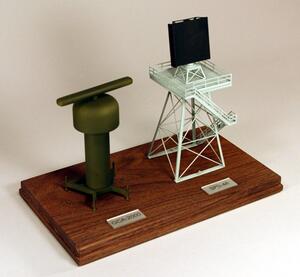 Two desktop radar models on a wooden base, angled view of an olive rotating antenna and a lattice tower with planar array