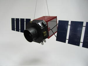 Generic satellite model hanging on wires, black telescope barrel facing left with red-wrapped body and blue solar arrays