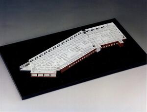 Roofless architectural model of an angled building showing white interior walls and red outer facade on a black base