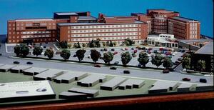 Hospital architectural model from a low angle, brick wings behind a tree-lined parking lot, with a plaque in foreground