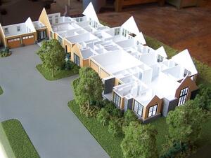 House model with roof removed, showing white interior walls and room layout, with driveway, garages and trees