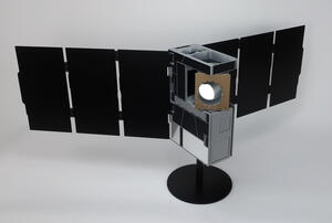 Sounders satellite model at three-quarter view with instrument aperture lit, solar arrays spread wide on a round black base