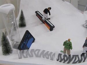 Snowboard display model from above, figure grinding a black rail beside a video screen, trees and spectator figures