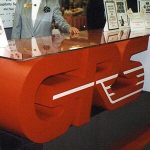 Trade show counter made of oversized red 3D logo letters with white stripes under a glass top, at an exhibit booth