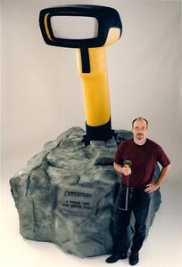 Giant barcode scanner model rising from a faux rock base, with a man holding a real handheld scanner for scale
