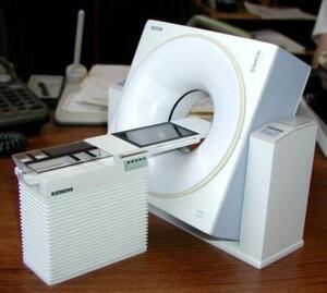 CT scanner scale model with ring gantry and patient table, shown at an angle on a wooden desk