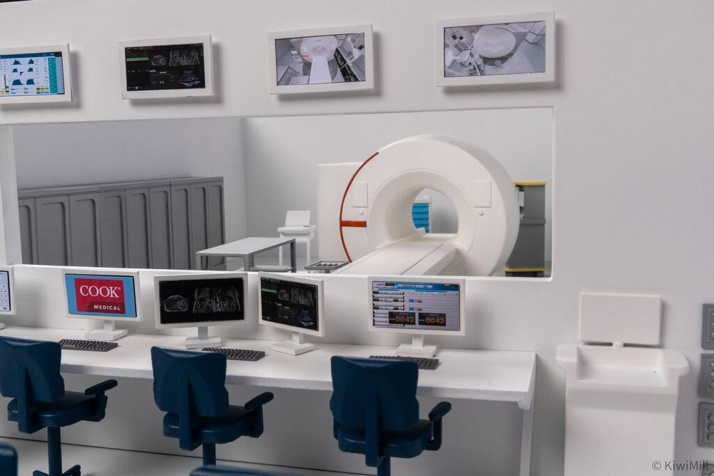 Control room desk with operator chairs and monitors, MRI scanner and storage cabinets visible through the window