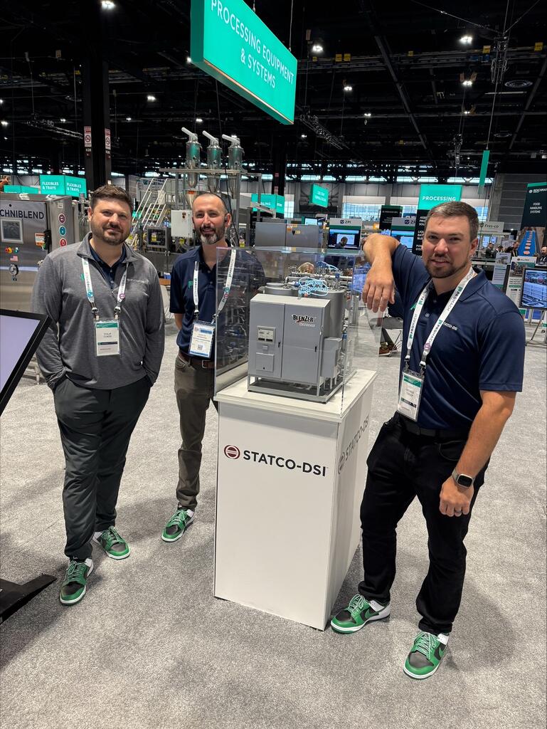BlenZer model in an acrylic case on a Statco-DSI pedestal at a trade show, with three booth staff beside it