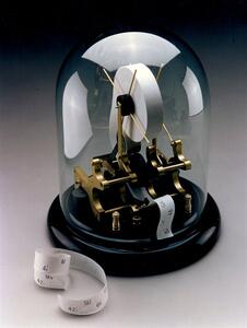 Ticker tape machine model under a glass dome on a black base, with brass mechanism and printed paper tape